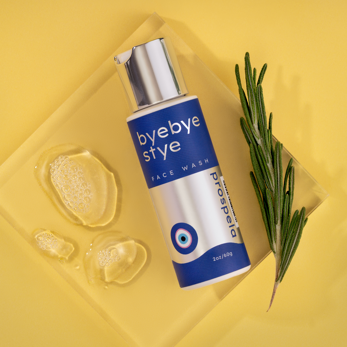 A bottle of bye bye stye face wash sits on a yellow background with a sprig of rosemary and gentle face wash suds. 
